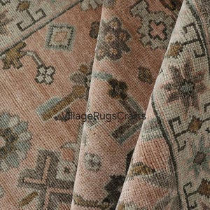May include: Close-up of a vintage rug with a distressed, faded appearance. The rug features a repeating geometric pattern in shades of dusty rose, grey, and brown. The rug is folded, showing the texture and detail of the weave.
