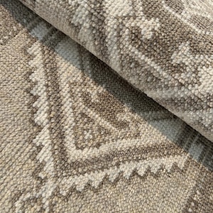 Pode incluir: Close-up de um tapete bege e castanho estampado. O tapete apresenta um design geométrico repetitivo com uma pilha texturizada e em laço. A paleta de cores inclui tons de bege, creme e castanho, criando uma estética neutra e versátil. O tapete está dobrado, revelando a parte inferior.