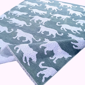 May include: A folded, teal-colored rug with a repeating pattern of white leopard silhouettes. The rug has a textured appearance and a visible edge, showcasing the fabric's weave.