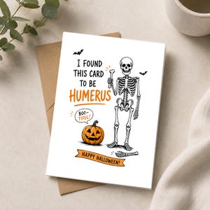 May include: A white greeting card featuring a black and white skeleton illustration holding a bone. The text reads I FOUND THIS CARD TO BE HUMERUS, BOO-TIFUL, and HAPPY HALLOWEEN. An orange jack-o'-lantern and two small black bats are also depicted.