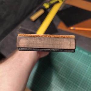 Puede incluir: Un bloque rectangular de madera con una parte superior de corcho y una base negra. El bloque se sostiene en la mano de una persona. El fondo incluye una regla amarilla, un l&aacute;piz y una esterilla de corte verde.
