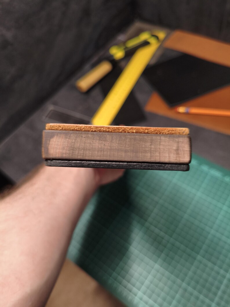 Puede incluir: Un bloque rectangular de madera con una parte superior de corcho y una base negra. El bloque se sostiene en la mano de una persona. El fondo incluye una regla amarilla, un l&aacute;piz y una esterilla de corte verde.