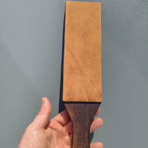 May include: A leather strop with a dark wooden handle. The rectangular leather surface is light brown, and the handle is a darker shade of brown. A small metal ring is attached to the bottom of the handle.