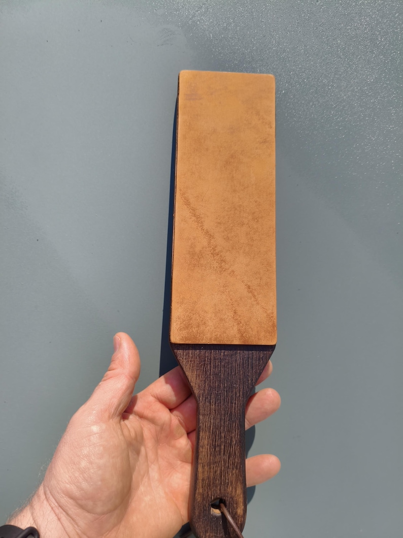 Puede incluir: Un asentador de cuero con un mango de madera oscura. La superficie rectangular de cuero es de color marr&oacute;n claro, y el mango es de un tono m&aacute;s oscuro de marr&oacute;n. Un peque&ntilde;o anillo de metal est&aacute; unido a la parte inferior del mango.