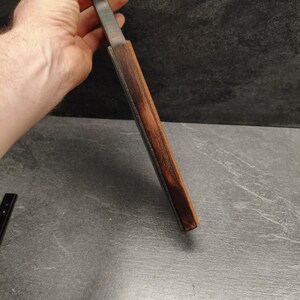 Puede incluir: Un cuchillo con una hoja de metal oscuro y un mango de madera. El mango est&aacute; envuelto en una funda de cuero marr&oacute;n. El cuchillo se sostiene en una mano sobre un fondo oscuro.