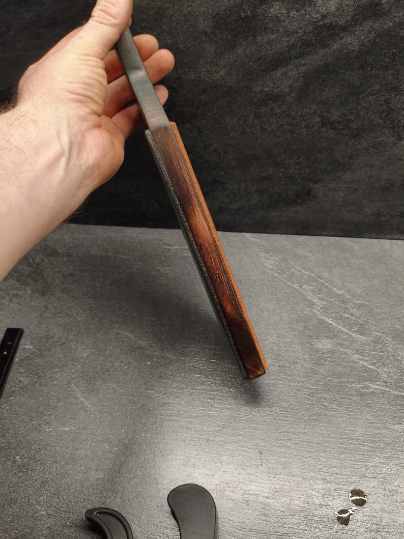 Puede incluir: Un cuchillo con una hoja de metal oscuro y un mango de madera. El mango est&aacute; envuelto en una funda de cuero marr&oacute;n. El cuchillo se sostiene en una mano sobre un fondo oscuro.