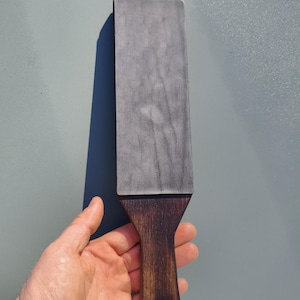 Puede incluir: Una piedra de afilar rectangular gris con un mango de madera oscura. El mango tiene un agujero para colgar y una correa de cuero. La piedra se sostiene sobre un fondo gris claro.