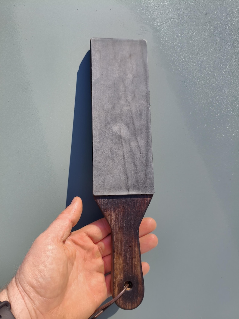 Puede incluir: Una piedra de afilar rectangular gris con un mango de madera oscura. El mango tiene un agujero para colgar y una correa de cuero. La piedra se sostiene sobre un fondo gris claro.