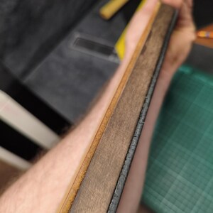 Puede incluir: Un cuero de madera y cuero, una herramienta para afilar navajas rectas. El cuero tiene una superficie de cuero marr&oacute;n claro y una superficie de madera m&aacute;s oscura. Los bordes son oscuros y la herramienta se sostiene en &aacute;ngulo.