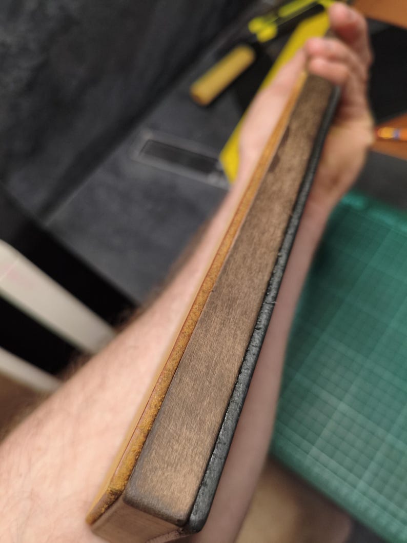 Puede incluir: Un cuero de madera y cuero, una herramienta para afilar navajas rectas. El cuero tiene una superficie de cuero marr&oacute;n claro y una superficie de madera m&aacute;s oscura. Los bordes son oscuros y la herramienta se sostiene en &aacute;ngulo.