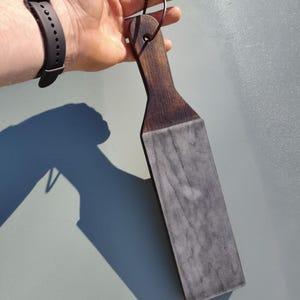 Puede incluir: Un asentador de madera oscura con una superficie de cuero gris. El asentador tiene un mango con un agujero para colgarlo y un lazo de cuero. La madera es de color marr&oacute;n oscuro y el cuero tiene un aspecto veteado.