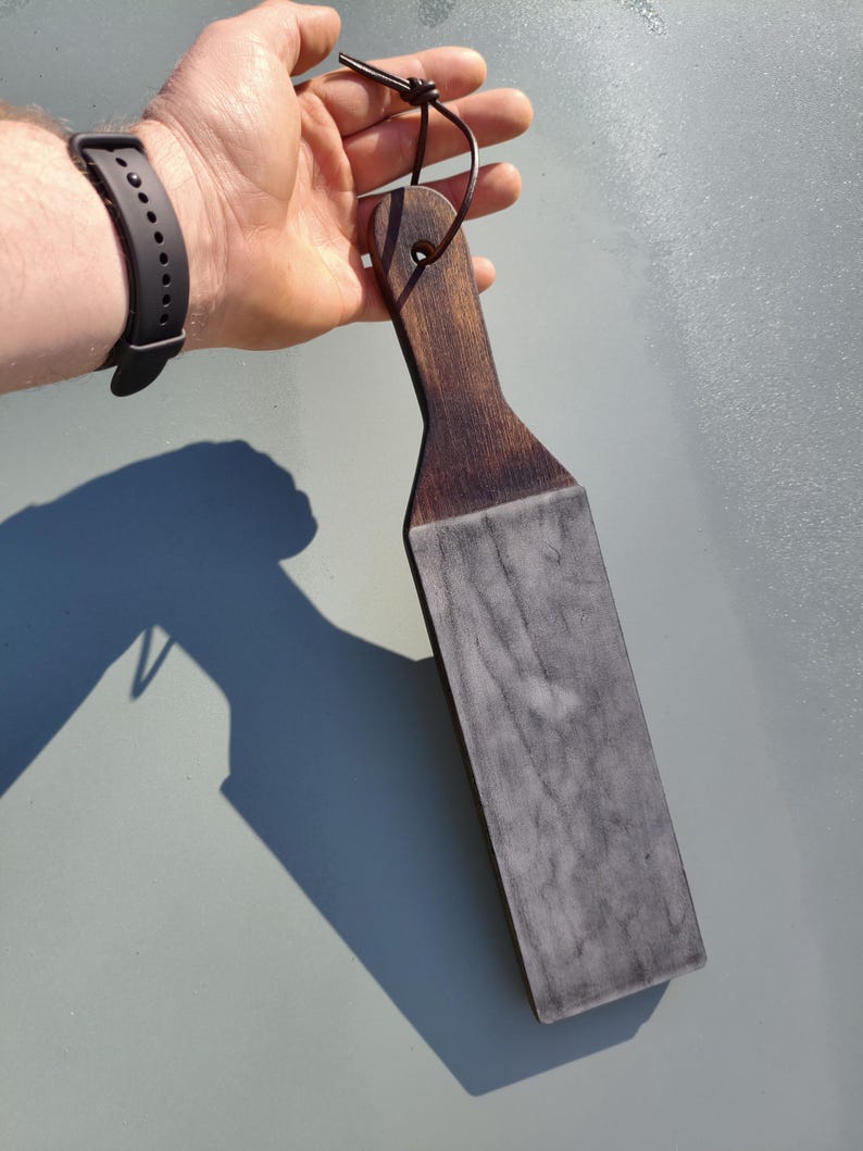 Puede incluir: Un asentador de madera oscura con una superficie de cuero gris. El asentador tiene un mango con un agujero para colgarlo y un lazo de cuero. La madera es de color marr&oacute;n oscuro y el cuero tiene un aspecto veteado.