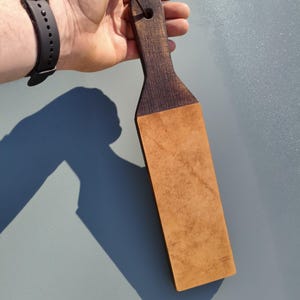 Puede incluir: Un asentador de madera con un mango marr&oacute;n oscuro y una superficie de cuero bronceado. El asentador se sujeta con un cord&oacute;n negro y est&aacute; dise&ntilde;ado para afilar navajas rectas. El mango tiene un agujero para colgar.