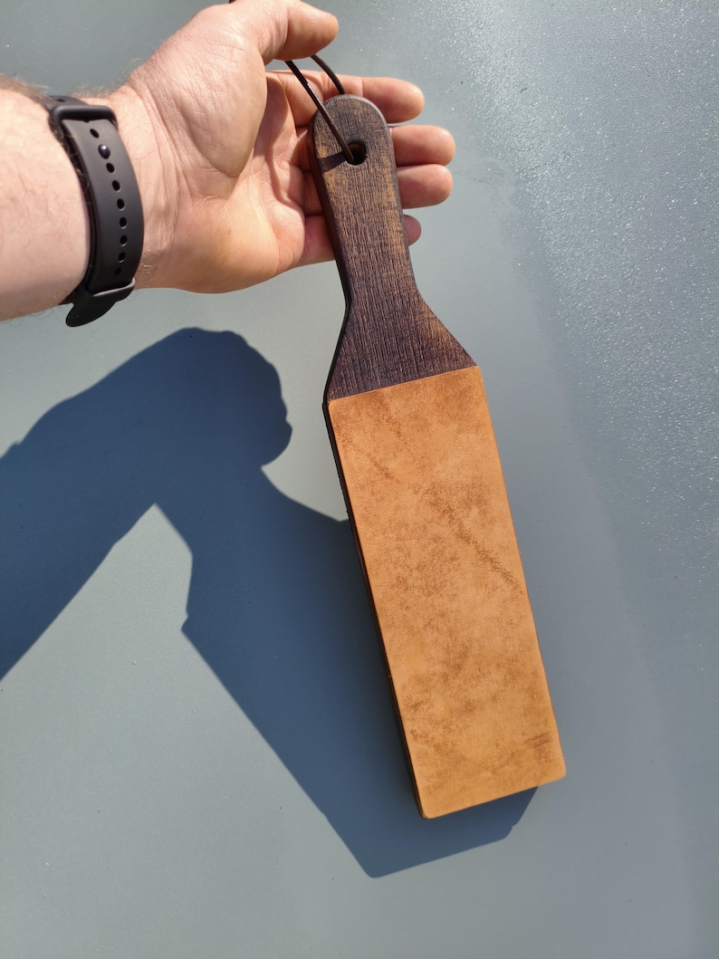 Puede incluir: Un asentador de madera con un mango marr&oacute;n oscuro y una superficie de cuero bronceado. El asentador se sujeta con un cord&oacute;n negro y est&aacute; dise&ntilde;ado para afilar navajas rectas. El mango tiene un agujero para colgar.
