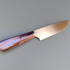 Peut inclure: Un couteau en acier inoxydable avec une lame polie et un manche présentant un motif tourbillonnant de violet, d'orange et d'or. La poignée a un point sombre et un anneau doré près de la lame.