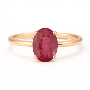 May include: A gold ring featuring an oval-cut ruby gemstone. The ruby is a deep red color and is held in place by four prongs. The band of the ring is a simple, polished gold.