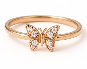 May include: A rose gold ring featuring a butterfly design. The butterfly has two wings with three round, clear gemstones on each wing. The ring band is thin and smooth. The ring is set against a white background.