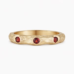 May include: A gold ring with a textured band and three round, red gemstones. The ring has a rustic, organic design, and the gemstones are evenly spaced along the band.