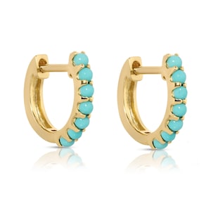 May include: A pair of gold-coloured hoop earrings. Each earring features a row of small, round, turquoise-coloured stones. The earrings are presented against a white background, with a subtle reflection beneath.