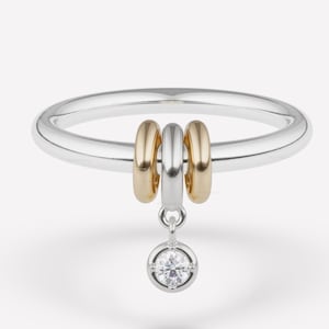 May include: A silver ring with three gold and silver accent rings. A small, round, clear gemstone hangs from the center of the ring. The ring is set against a white background.