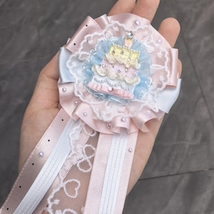 May include: A circular rosette badge featuring a central layered cake motif in pastel yellow, pink, and blue. The badge is adorned with white lace, satin ribbons in pale pink and white, small faux pearls, and clear rhinestones on the cake layers.