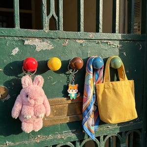 Könnte beinhalten: Eine Sammlung von Gegenständen, die an einer grünen Tür hängen. Dazu gehören ein rosa Plüschhase, ein Schlüsselanhänger mit einem Zeichentier, ein gemusterter Schal und eine gelbe Tragetasche. Die Tür hat dekorative Metallarbeiten und bunte Knöpfe.