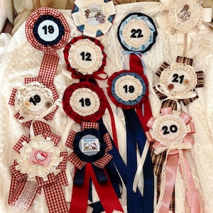 May include: A collection of circular fabric rosettes featuring lace trim, ribbons, and various numbers including 12, 18, 19, 20, 21, and 22. Some rosettes display Hello Kitty graphics, plaid patterns, and solid colors like red, blue, and pink.