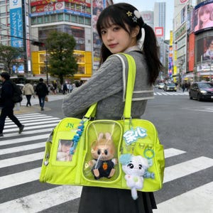 May include: A bright neon green tote bag with a clear pocket containing a plush bunny wearing a suit. The bag has a photo, a blue beaded charm, and a white and blue plush dog. The bag is carried on the shoulder.