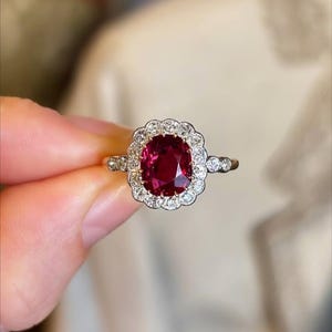 Peut inclure: Une bague en or ornée d'une grosse pierre rouge foncé de taille ovale, entourée d'un halo de plus petites pierres précieuses claires. La monture de la bague est également ornée de petites pierres précieuses claires de chaque côté de la pierre centrale. La bague est tenue par une main.