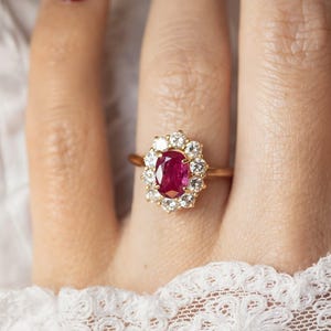 Peut inclure: Bague en or ornée d'un rubis ovale central entouré d'un halo de diamants. La monture en or fin met en valeur le rouge vif du rubis et l'éclat des diamants. La bague est portée sur un doigt avec du vernis à ongles rouge.