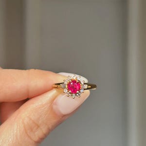Può includere: Un anello d'oro con una vivace pietra rossa al centro, circondata da un alone di pietre più piccole e trasparenti. La fascia dell'anello è sottile e l'incastonatura mostra un design classico ed elegante. L'anello è tenuto da una mano.