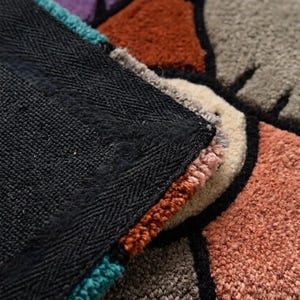 Puede incluir: Una alfombra de mechones con un patr&oacute;n geom&eacute;trico de bloques en morado, gris, naranja &oacute;xido y crema, delineados con hilo negro grueso. Se observa el reverso de la alfombra, que muestra un forro de tela negra s&oacute;lida con textura tejida.