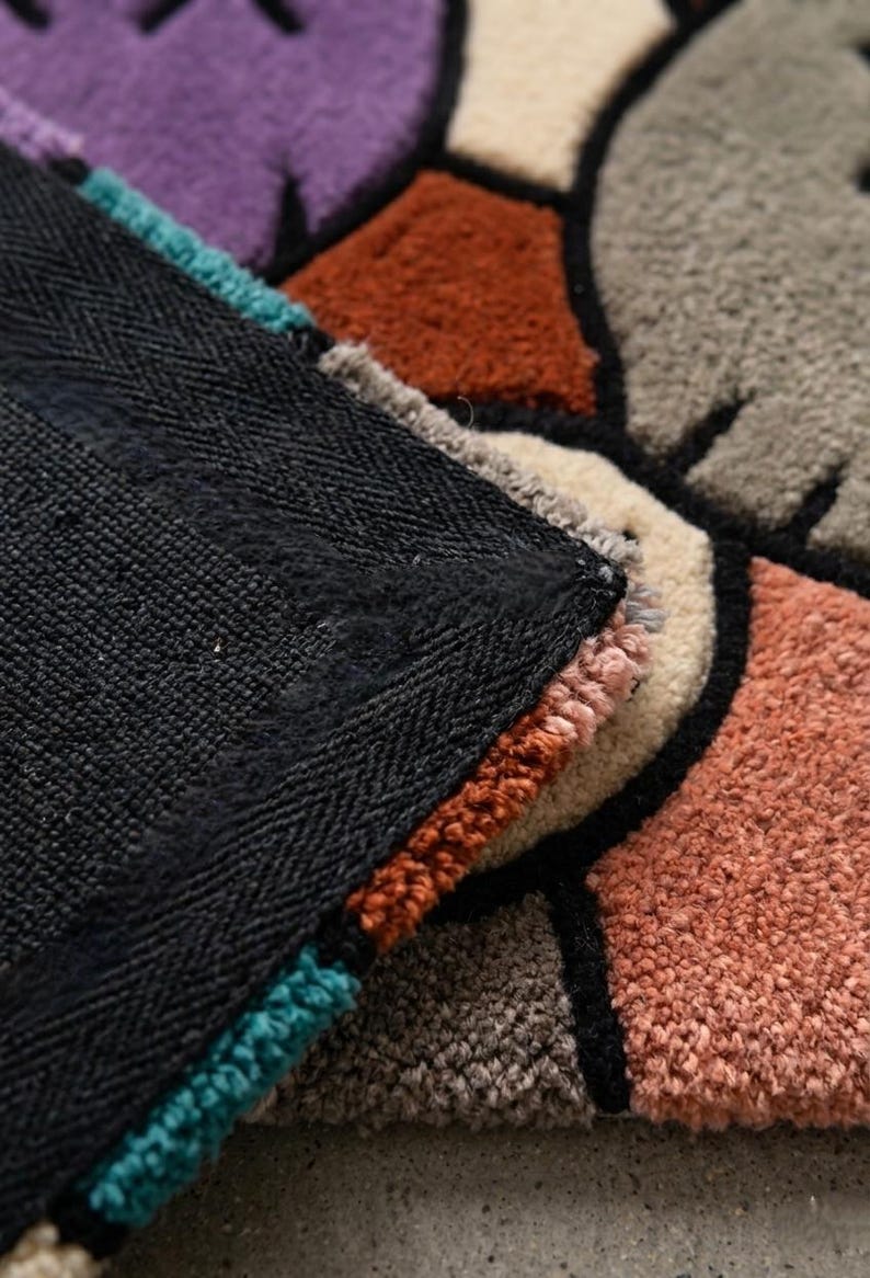 Puede incluir: Una alfombra de mechones con un patr&oacute;n geom&eacute;trico de bloques en morado, gris, naranja &oacute;xido y crema, delineados con hilo negro grueso. Se observa el reverso de la alfombra, que muestra un forro de tela negra s&oacute;lida con textura tejida.