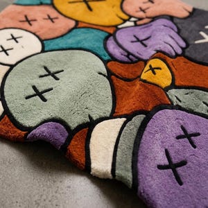 Puede incluir: Una alfombra de felpa con un vibrante patr&oacute;n abstracto multicolor de formas redondeadas. Cada forma contiene un s&iacute;mbolo de 'X' negro en lugar de ojos. La paleta de colores incluye tonos amarillos, morados, verde azulado, naranjas, verdes y crema, todos delineados en negro.