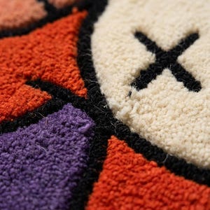 Puede incluir: Una alfombra de mechones con una superficie texturizada y bloques de colores geom&eacute;tricos en naranja, morado y crema. El dise&ntilde;o incluye una forma circular de color crema marcada con una X negra cosida, todo delineado con hilo negro grueso.