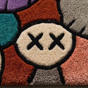 Puede incluir: Una alfombra de mechones con un patr&oacute;n geom&eacute;trico multicolor con varias formas en morado, verde azulado, naranja &oacute;xido, beige, gris y rosa. Una forma circular beige central muestra dos s&iacute;mbolos X negros cosidos, con todas las secciones delineadas en negro.