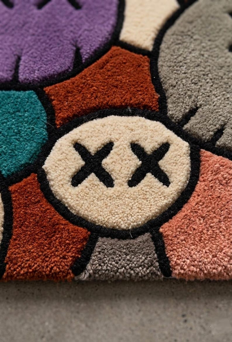 Puede incluir: Una alfombra de mechones con un patr&oacute;n geom&eacute;trico multicolor con varias formas en morado, verde azulado, naranja &oacute;xido, beige, gris y rosa. Una forma circular beige central muestra dos s&iacute;mbolos X negros cosidos, con todas las secciones delineadas en negro.