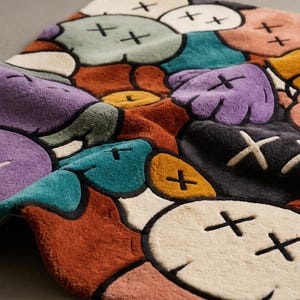 Puede incluir: Una alfombra rectangular con un patr&oacute;n multicolor de formas redondeadas similares a nubes. Cada forma contiene dos marcas de X negras bordadas como ojos. La paleta de colores incluye tonos crema, verde azulado, morado, naranja y gris oscuro, todo delineado en negro.
