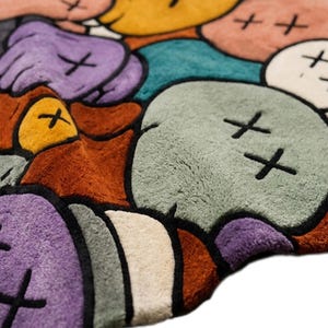 Puede incluir: Una alfombra de felpa con un patr&oacute;n repetitivo de cabezas de personajes redondeadas tipo dibujos animados en tonos amarillo, morado, verde azulado, verde claro, naranja y crema. Cada cabeza est&aacute; delineada en negro con dos X negras.