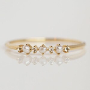 May include: A delicate gold ring featuring a row of small, round pearls and sparkling accent stones. The ring's band is thin and polished, with the pearls and stones evenly spaced across the top. The ring is set against a white background.