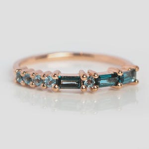 May include: A rose gold ring featuring a row of alternating blue-green baguette and round-cut gemstones. The ring's band is a polished rose gold, and the gemstones are set in a channel setting.