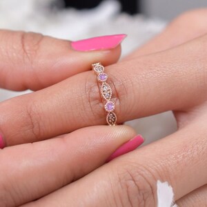 May include: A rose gold ring with a repeating pattern of oval and round gemstones. The ring features alternating pink and clear stones, with intricate detailing along the band. The ring is held between two hands with pink nail polish.