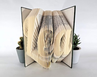 Könnte beinhalten: Eine Buchkunstskulptur mit den gefalteten Seiten eines Hardcover-Buches, die die Buchstaben L-E-O bilden. Das Buch hat einen dunkelgrünen Einband und steht zwischen zwei kleinen Sukkulenten in grauen und weißen Keramik-Pflanzgefäßen.