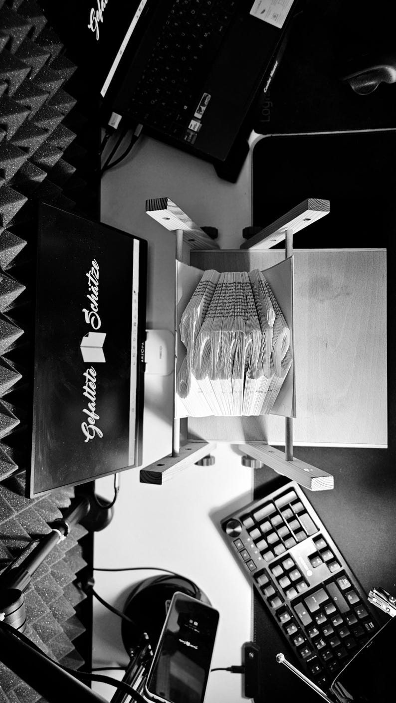 May include: A black and white overhead view of a book art sculpture featuring the name Jamie folded into the pages. The sculpture is held in a wooden stand next to a computer monitor displaying the text Gefaltete Sch&auml;tze and a keyboard.