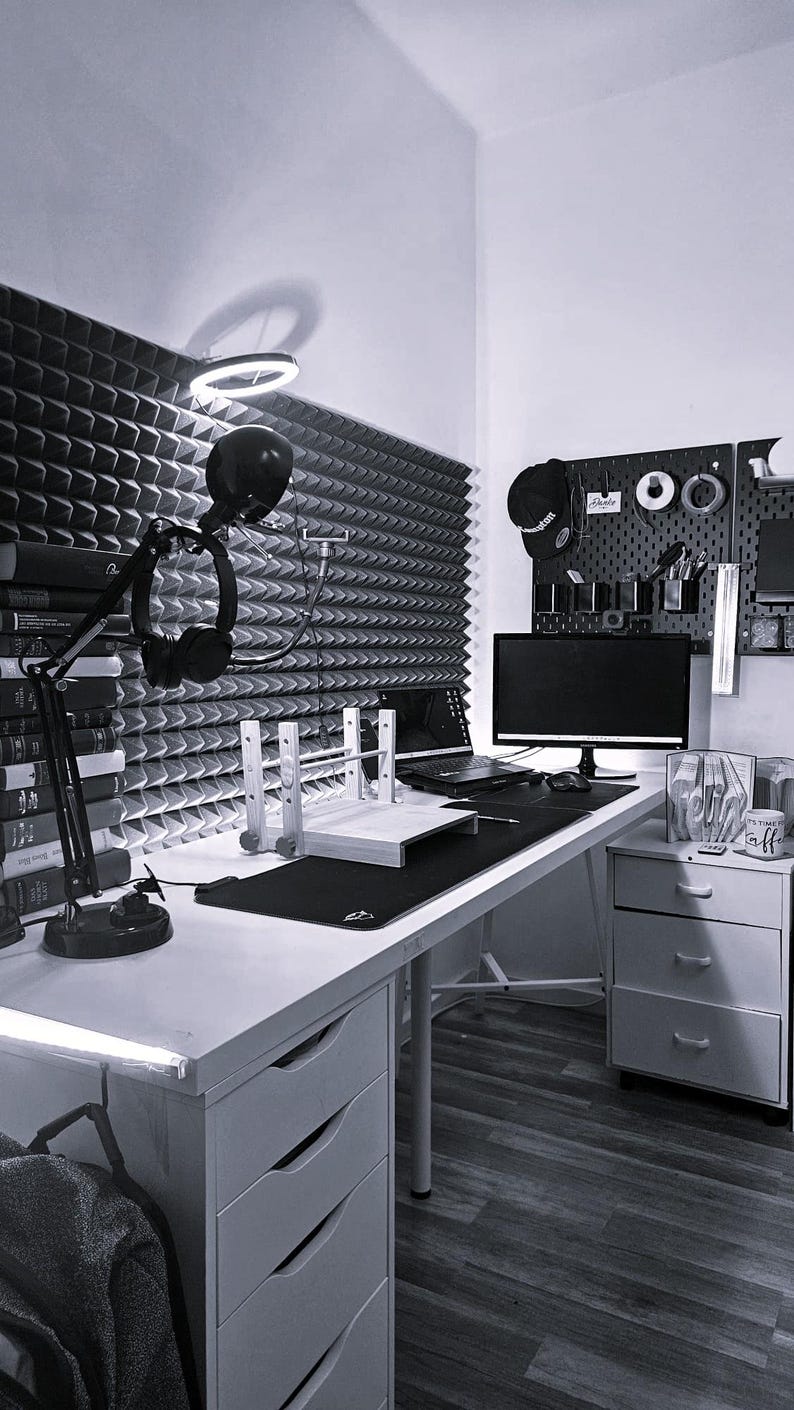 May include: A home office setup featuring a white desk with a black desk mat, a computer monitor, a laptop, and a microphone on an adjustable arm. The wall is covered in grey acoustic foam panels, and a black pegboard holds various accessories.