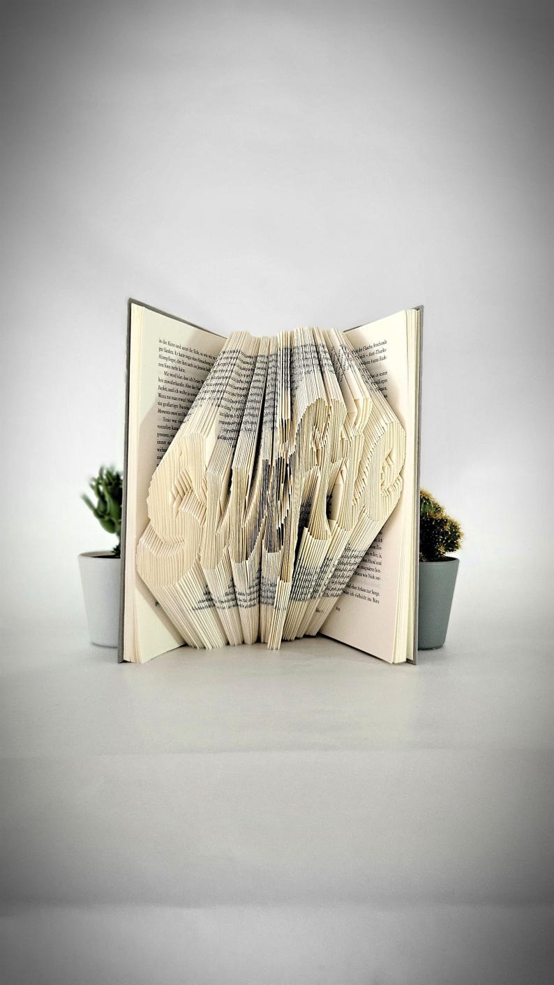 May include: A book with pages folded to form the word Swift, displayed in an open position. The paper pages show printed black text, and the book is positioned between two small potted green plants with grey containers on a plain surface.