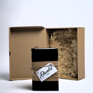May include: A black notebook wrapped in a brown paper band with twine and a white tag featuring the word Danke in black script. The notebook is positioned in front of an open brown cardboard gift box filled with light-coloured shredded paper filler.