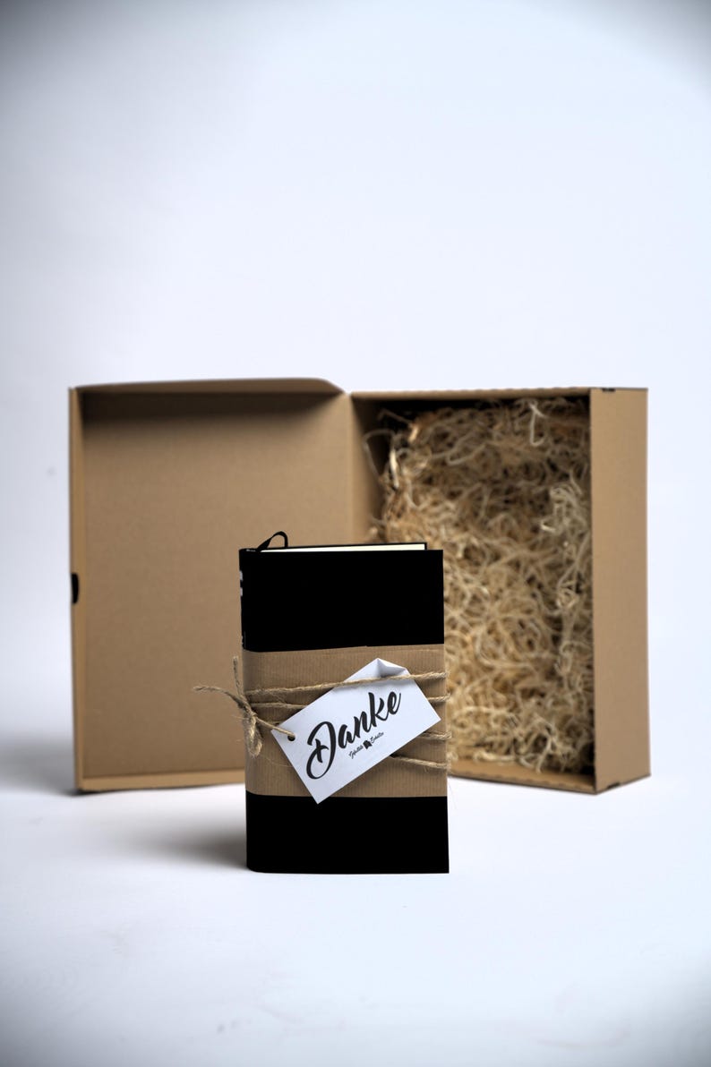 May include: A black notebook wrapped in a brown paper band with twine and a white tag featuring the word Danke in black script. The notebook is positioned in front of an open brown cardboard gift box filled with light-coloured shredded paper filler.