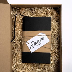 May include: A black notebook with a black ribbon bookmark, wrapped in a brown paper band and tied with twine. A white rectangular tag with the word Danke in black script is attached to the twine. The notebook rests inside a cardboard box.