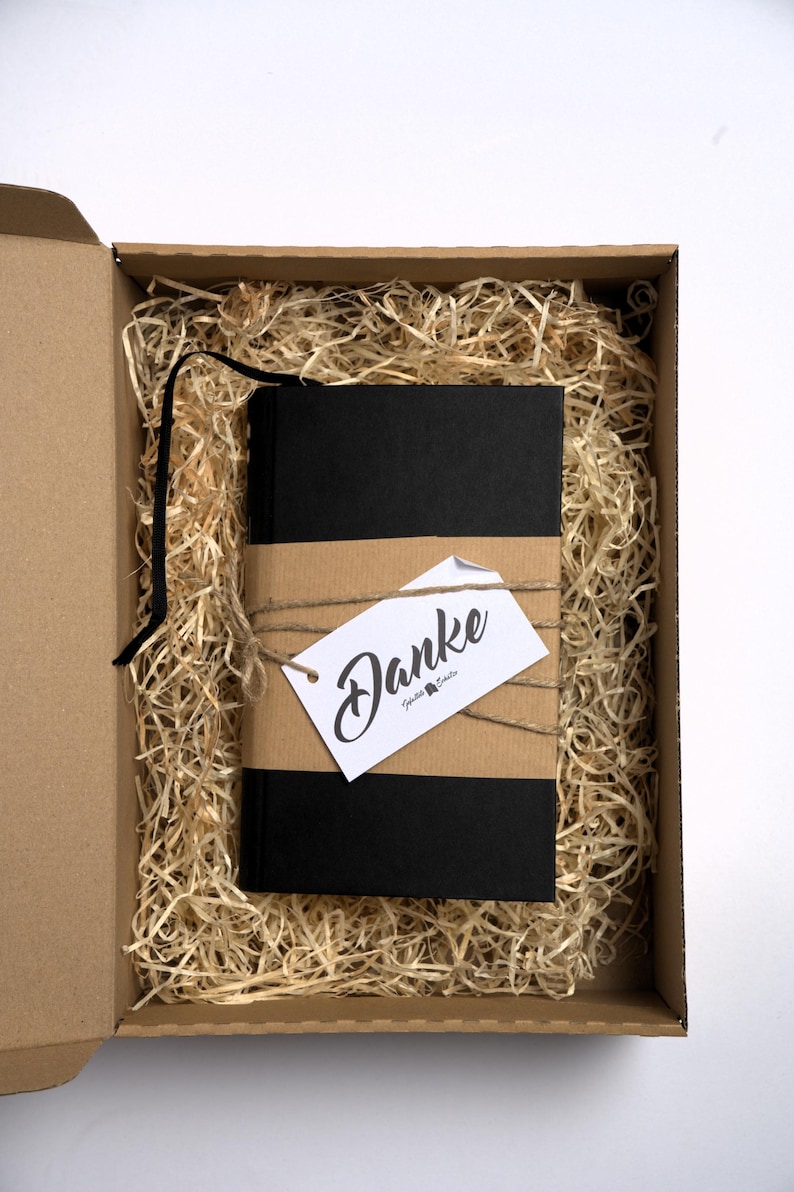 May include: A black notebook with a black ribbon bookmark, wrapped in a brown paper band and tied with twine. A white rectangular tag with the word Danke in black script is attached to the twine. The notebook rests inside a cardboard box.